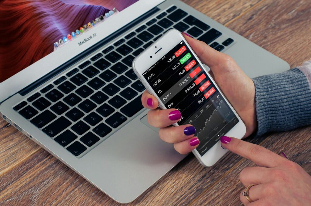 A person uses a smartphone displaying a stock market app in front of an open MacBook Air on a wooden desk, researching the next valuable asset—be it shares or a house.