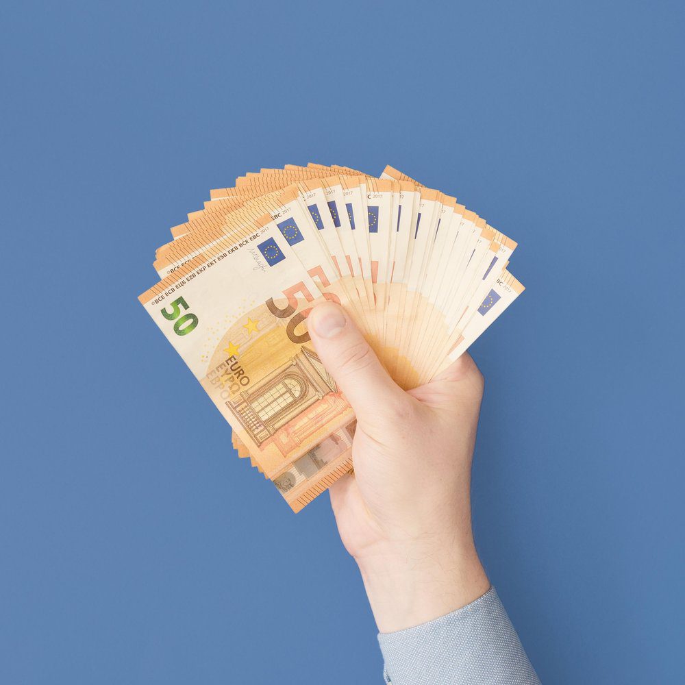 A hand holding a fanned-out stack of fifty euro banknotes against a plain blue background, symbolizing unclaimed mortgage relief.
