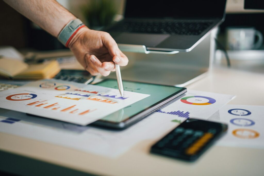 Person pointing at printed charts on a tablet with a pen, surrounded by graphs, a smartphone, and a laptop on a desk—analyzing rates and recent ECB data in a neutral setting.
