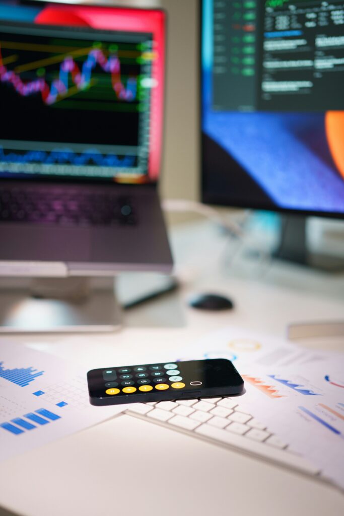 A smartphone with a trusted calculator app is placed on printed financial charts, with a keyboard, laptop, and monitor displaying graphs and data in the background.