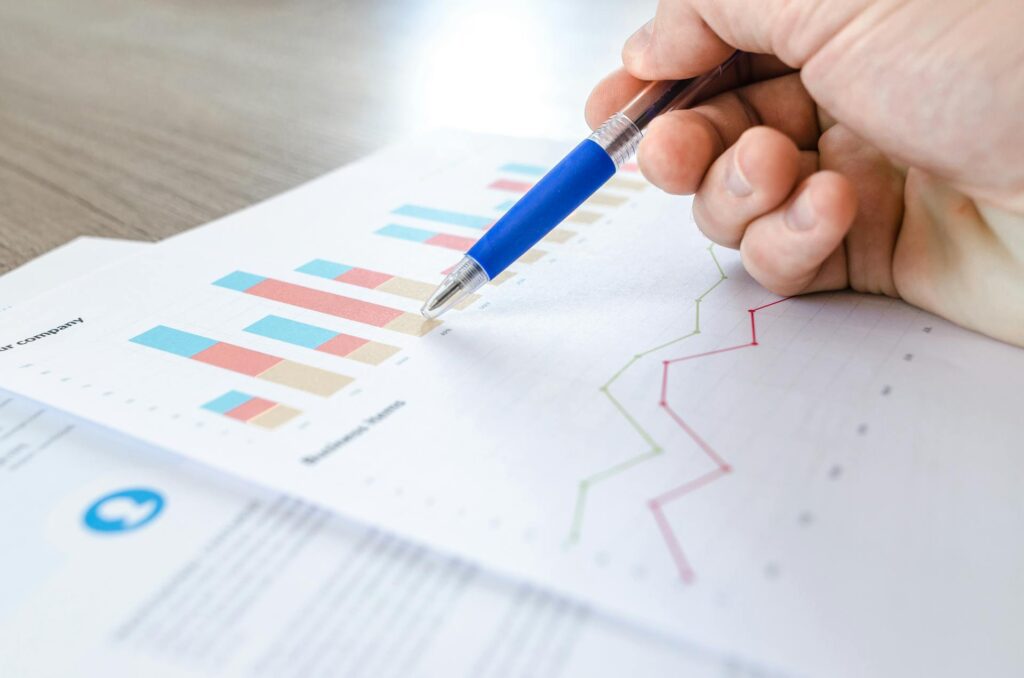 A hand holding a pen points to a bar chart on a printed paper with colorful graphs and data, encouraging you to start thinking like a No.1 analyst, all placed on a desk.