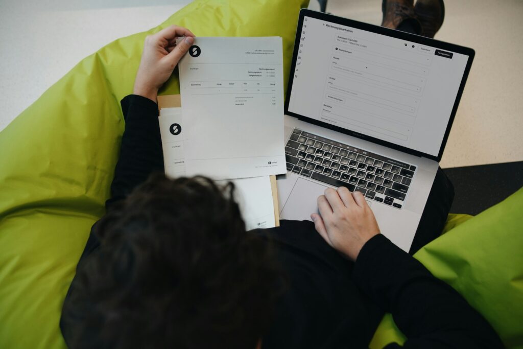 Person sitting on a green beanbag, holding documents in one hand and easily filling out an online form on their laptop.