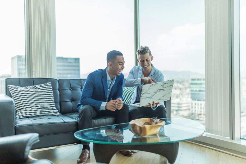 Two men in business attire sit on a couch in a modern office, smiling as they review planning strategies on a laptop, with city buildings visible through large windows behind them.