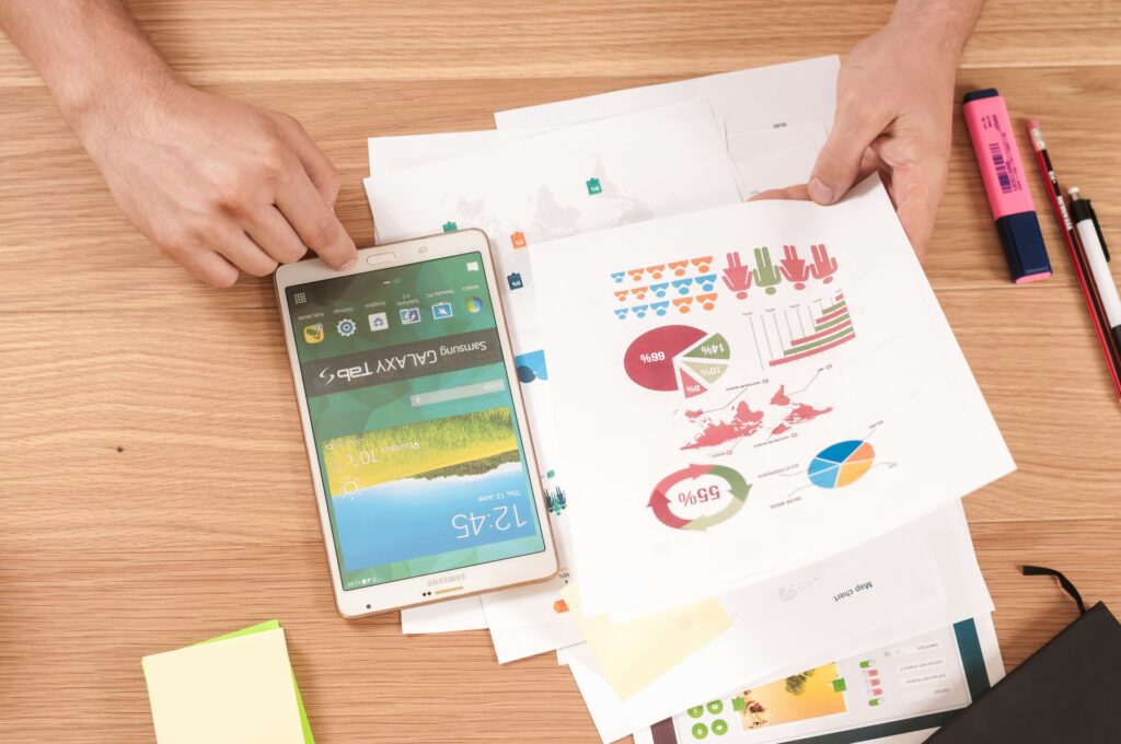 A person reviews printed charts and graphs on paper next to a Samsung tablet, highlighters, pens, and a handwritten Christmas message on a wooden desk.