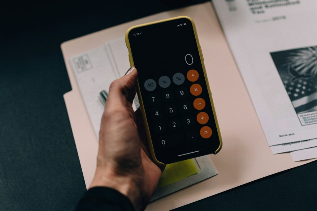 A person holds a smartphone with a calculator app open, displaying zero, over a desk with documents and a folder—ideal for planning and tracking financial goals.