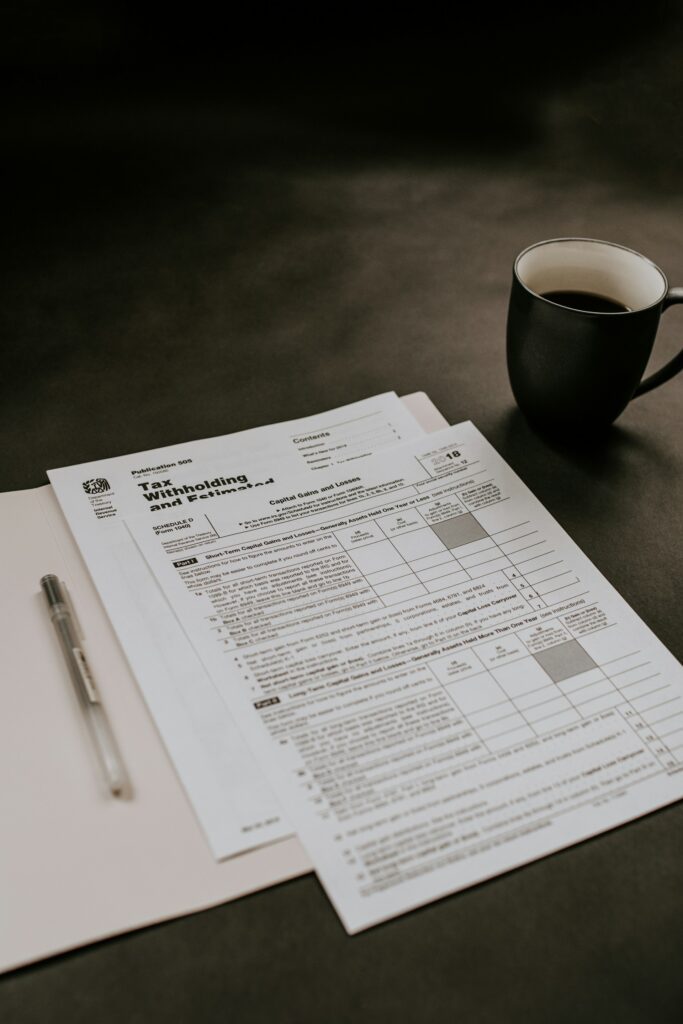 A stack of tax forms, a pen, a folder, and a cup of black coffee on a dark tabletop captures the contrast between the have vs have not during tax season.