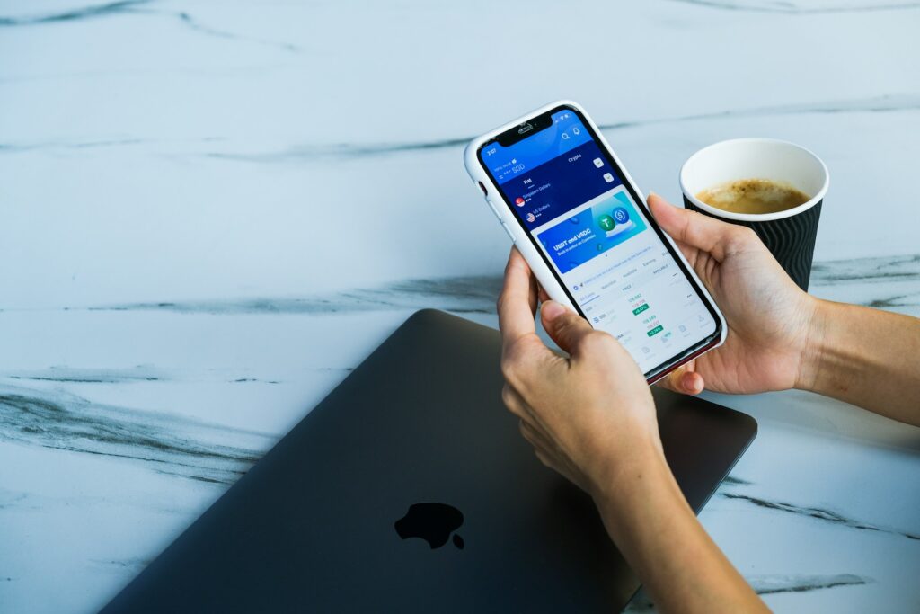 Person holding a smartphone displaying a financial app focused on retirement planning, with a closed MacBook and a cup of coffee on a white marble surface.
