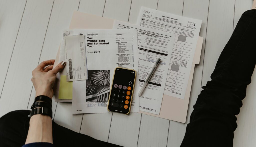 Person sitting on the floor with tax documents, a pen, a phone with a calculator app open, and an envelope on top of a folder—ready to create their own client success stories.