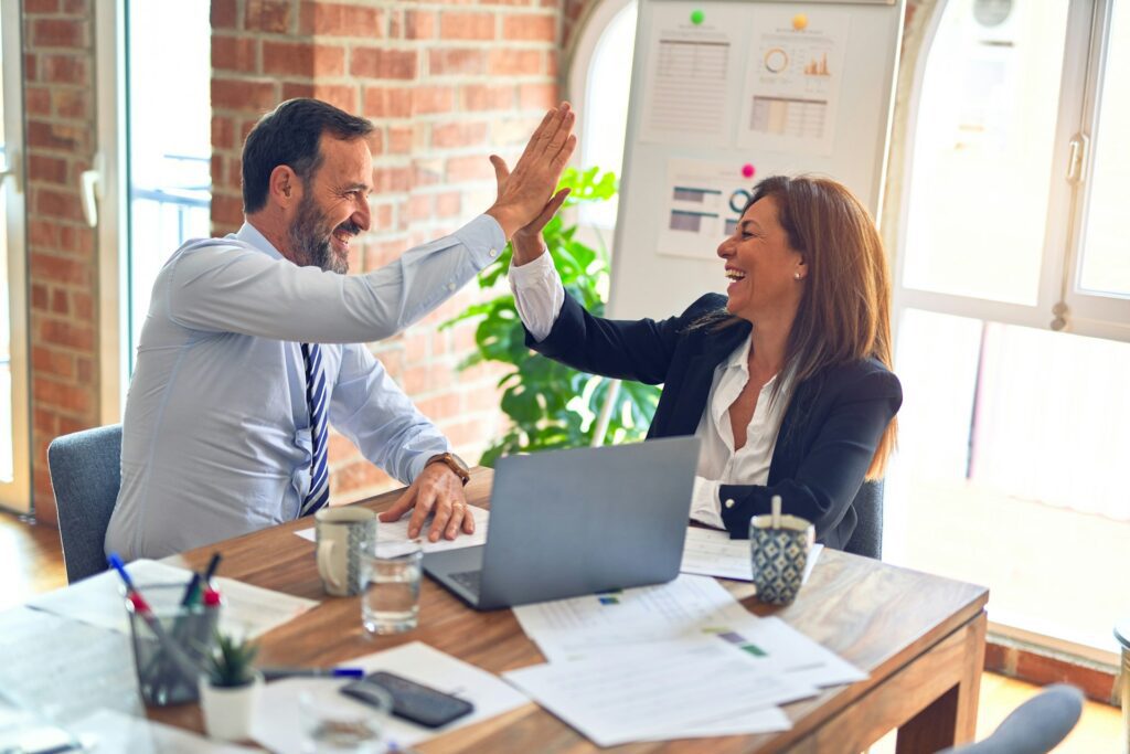Two colleagues sit at a desk, smiling and giving each other a high five after sharing a great mortgage tip, with papers and a laptop in front of them in a bright office setting.