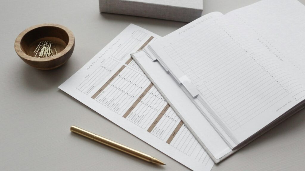 A gold pen, finance tracker sheets, an open planner, and a wooden bowl with paperclips are arranged on a light desk—perfect for organizing credit card bills.
