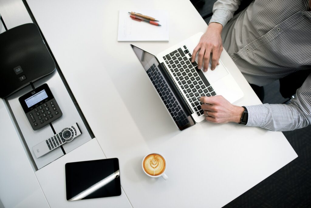 Person typing on a laptop at a white desk with coffee, a notepad and pens, phone, and tablet—perfect for staying organized while focusing on pension management or retirement planning.
