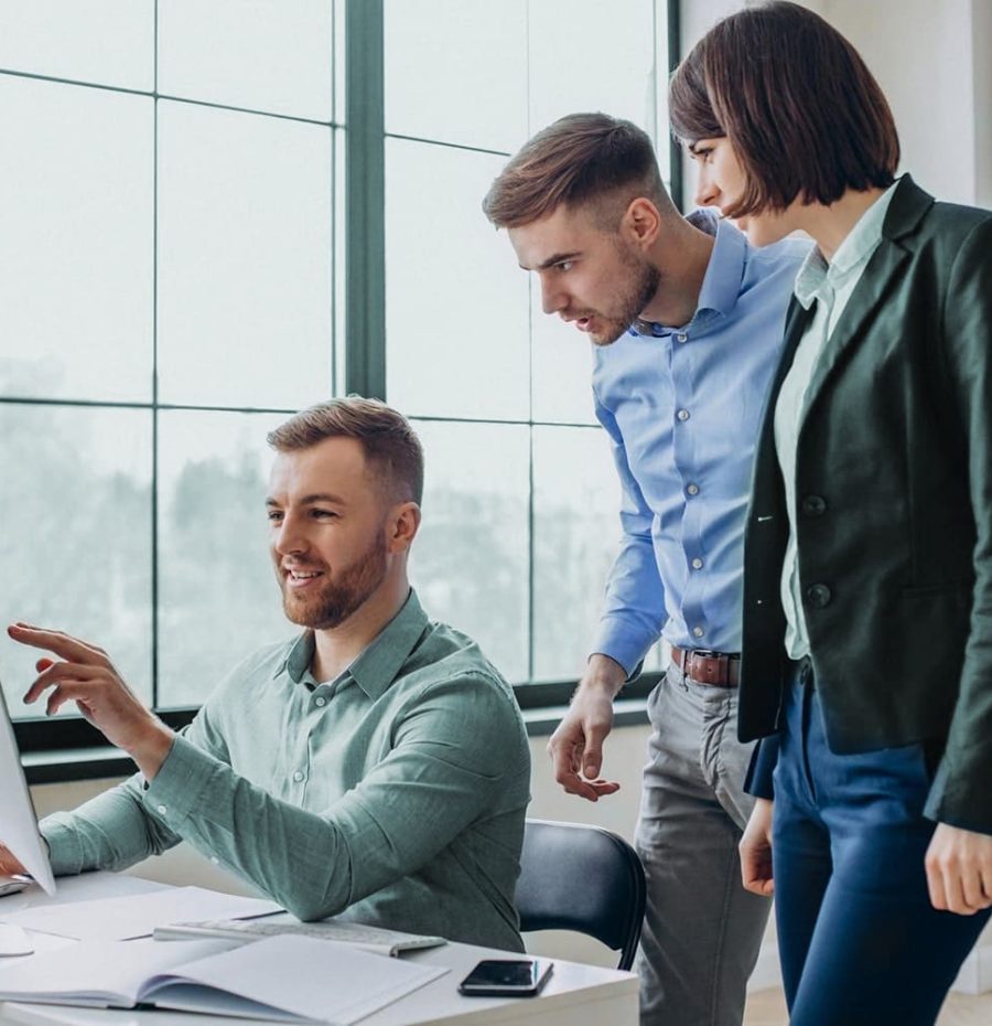 Three people in business attire look at a laptop together in a bright office, with one person seated and pointing at the screen while the others stand nearby.