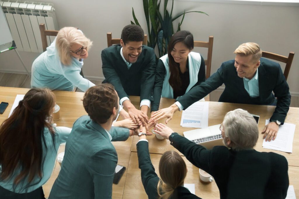 A group of eight people in business attire sit around a table, placing their hands together in the center as a gesture of teamwork.