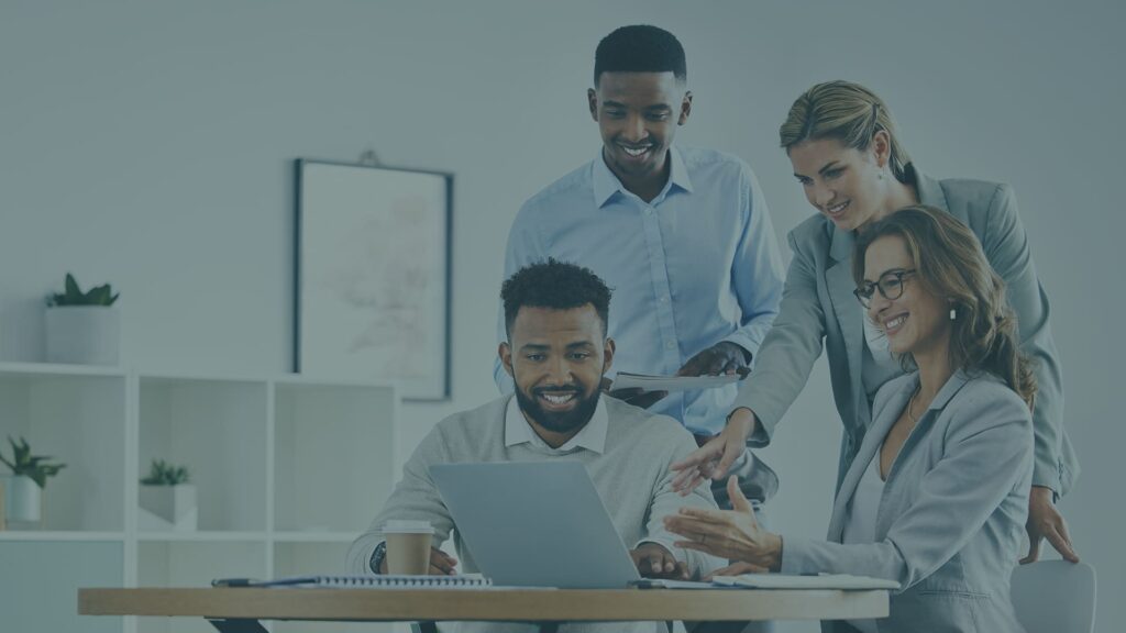 Four professionals collaborate around a laptop at a table in an office setting, appearing engaged and focused on their work.