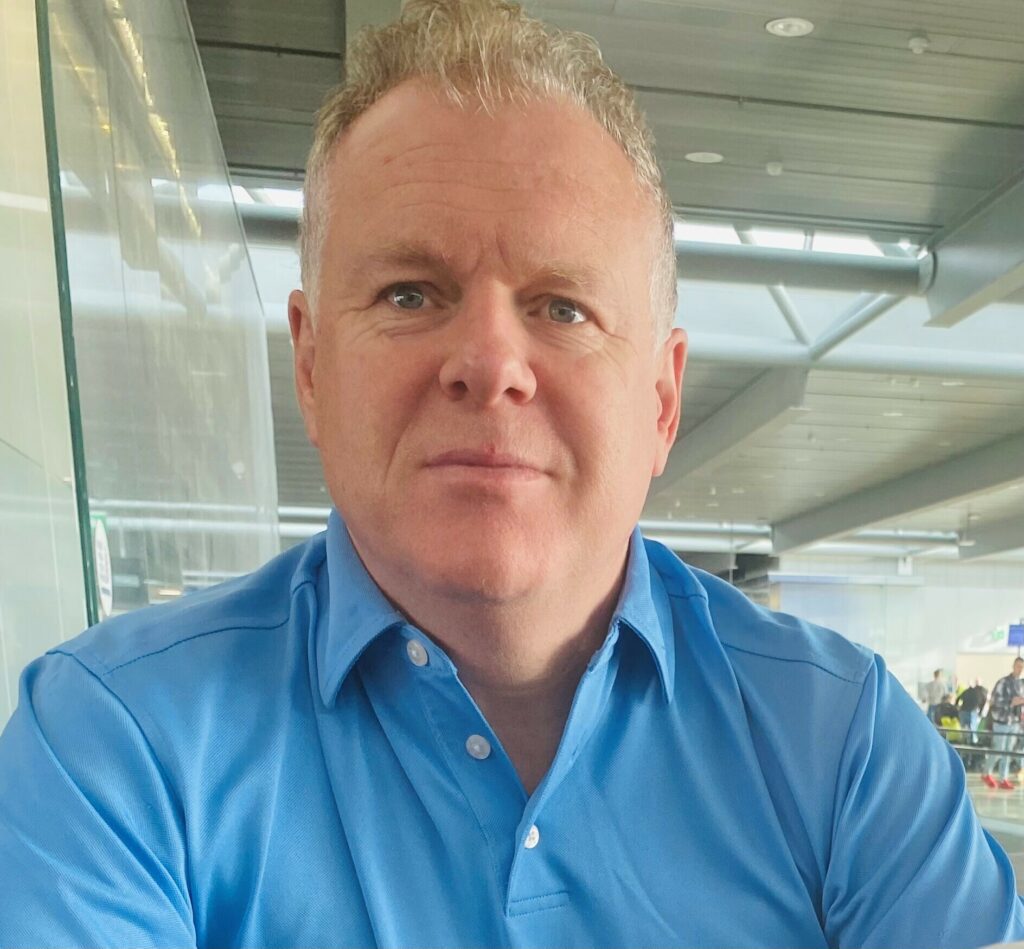 A man with short, curly hair wearing a light blue collared shirt sits indoors in a modern, well-lit building.