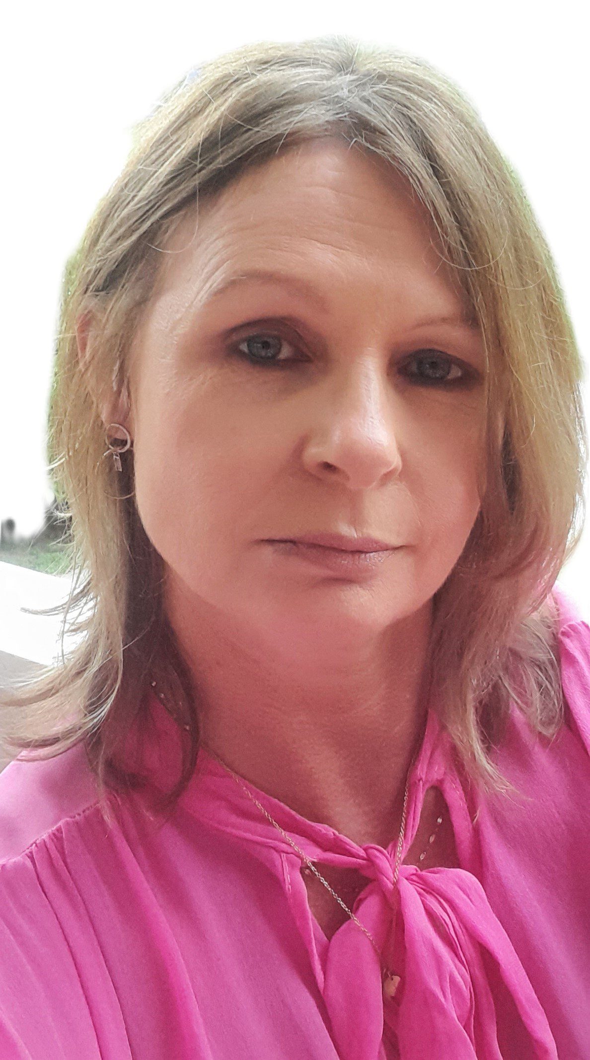 A person with shoulder-length blonde hair wears a pink blouse and looks at the camera with a neutral expression. The background is out of focus.