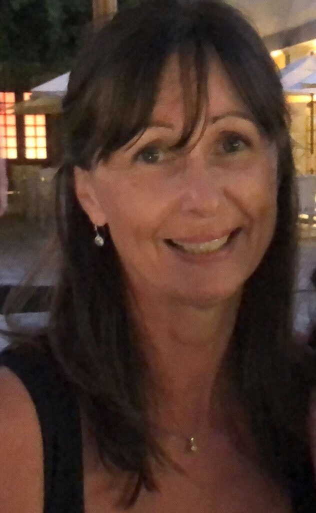 A woman with long brown hair and a necklace smiles at the camera; outdoor setting with blurred lights and umbrellas in the background.