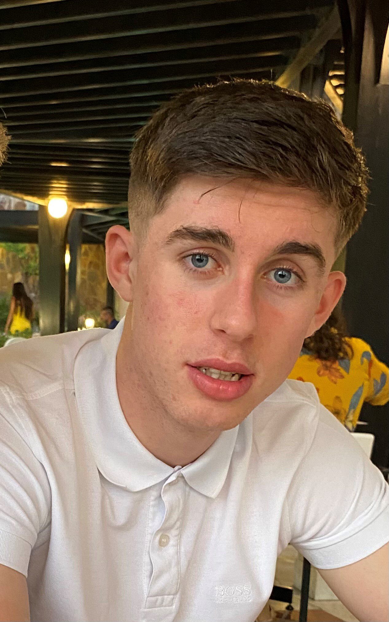 A young man with short brown hair and blue eyes wearing a white polo shirt sits indoors, looking at the camera.