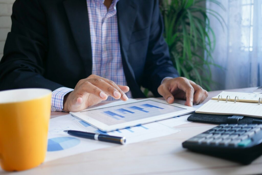 A business professional in a suit reviews bar charts on a tablet at a desk with documents, a notebook, a pen, a calculator, and a yellow cup—analyzing data like carefully playing Jenga.