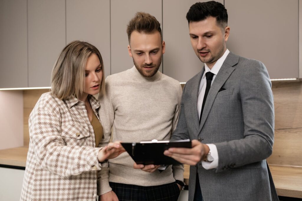 Three people stand together indoors, reviewing documents on a clipboard; one man wears a suit, while the other man and woman are dressed casually.