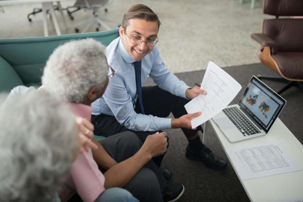 A man in business attire offers business advice as he shows documents to two older adults while sitting on a couch, with a laptop displaying a colorful chart on the table.