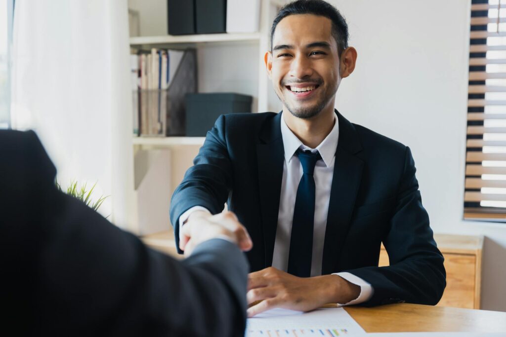 A man in a suit smiles while shaking hands with another person across a desk, celebrating a new partnership at Raisin Bank in an office setting.