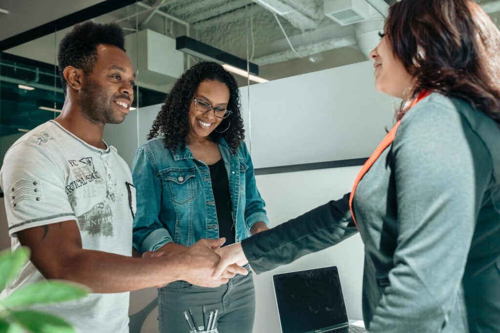 Two people stand and smile as one of them shakes hands with a woman in professional attire across a desk, discussing services in an office setting.