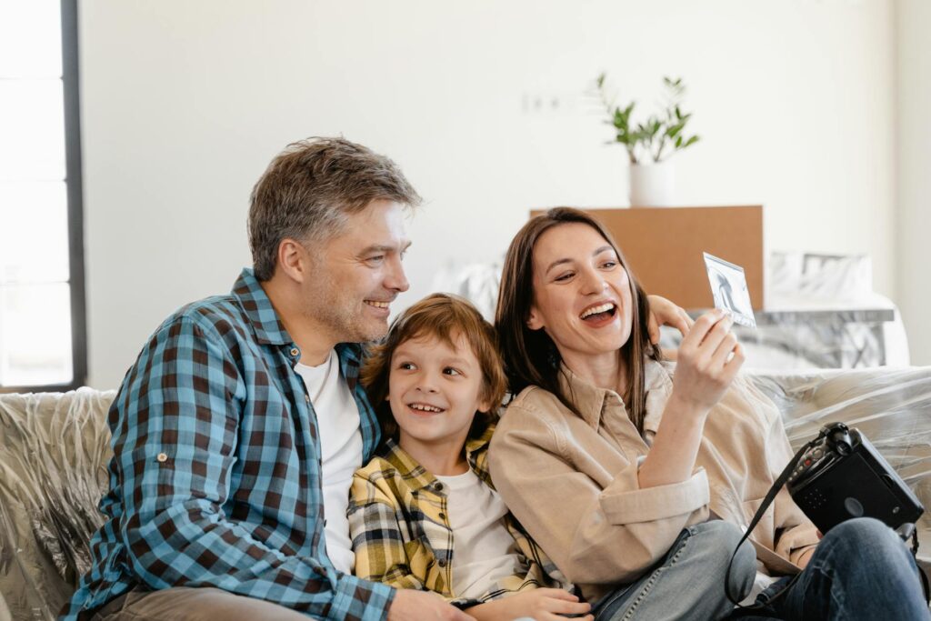 A smiling man, woman, and young boy sit together on a couch, holding and looking at a photo, with moving boxes and a plant in the background—celebrating new beginnings made possible by their savings.