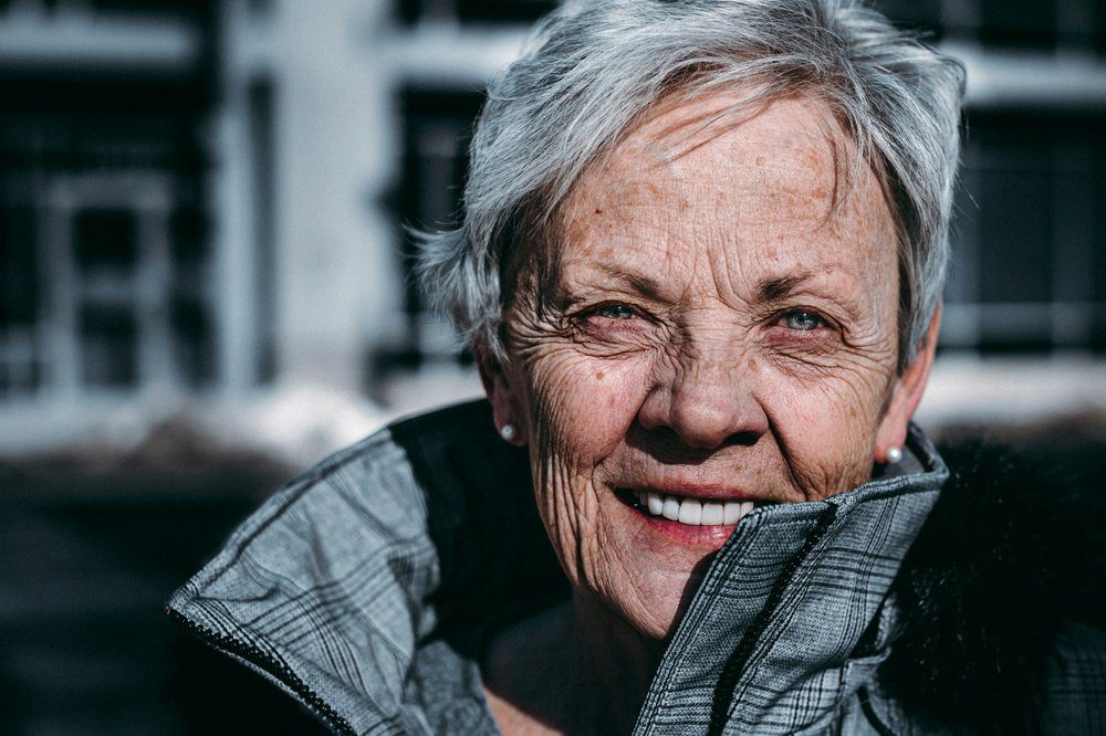 Older woman with short gray hair smiles outdoors, wearing a light gray jacket. The background is blurred and dark, highlighting her face in bright sunlight as she enjoys using Planning Calculators to organize her next adventure.