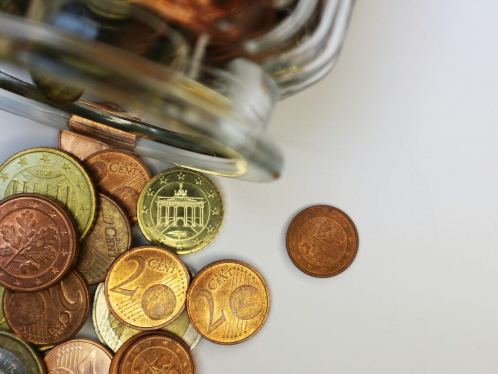 A glass jar tipped over with euro coins spilling onto a white surface, including 1-euro, 20-cent, and other euro cent coins.